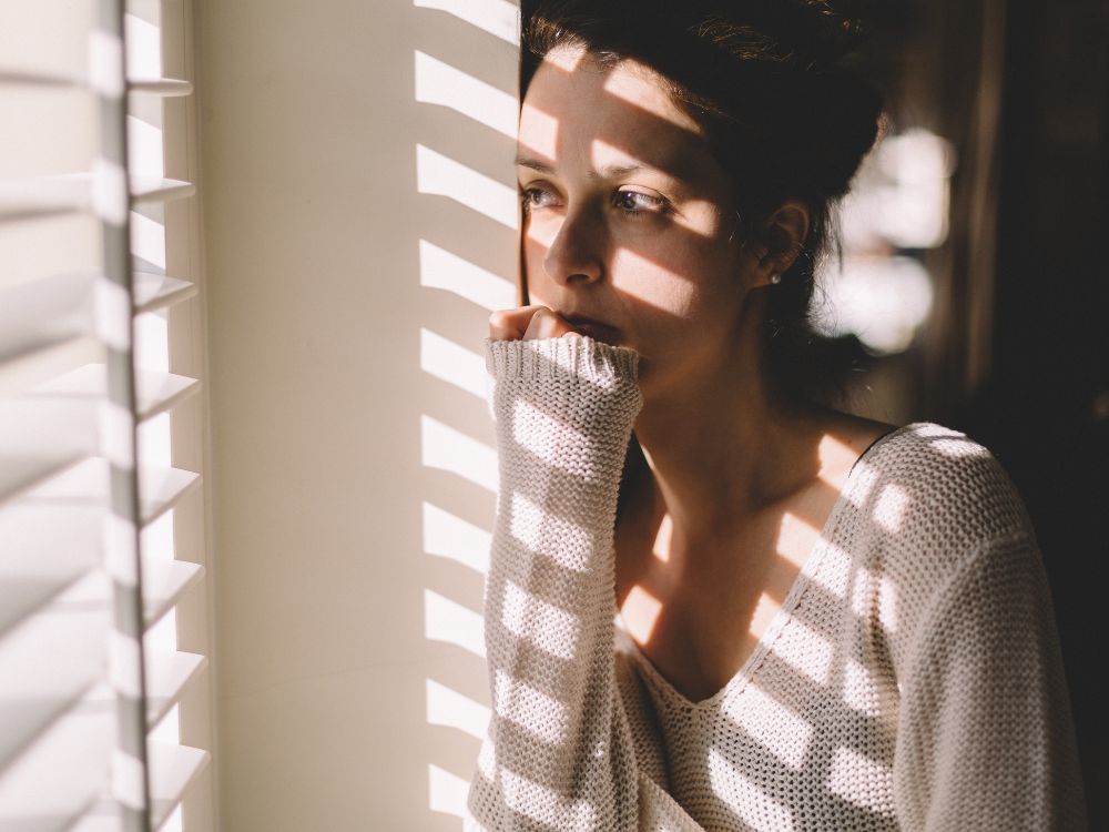 Cuando la T4 no es suficiente Deficiencia de T3 y enfermedad de la tiroides Parte 1: Imagen de una mujer melonchólica con un suéter mirando por la ventana.